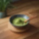 A vibrant bowl of matcha powder next to a bamboo whisk on a wooden surface